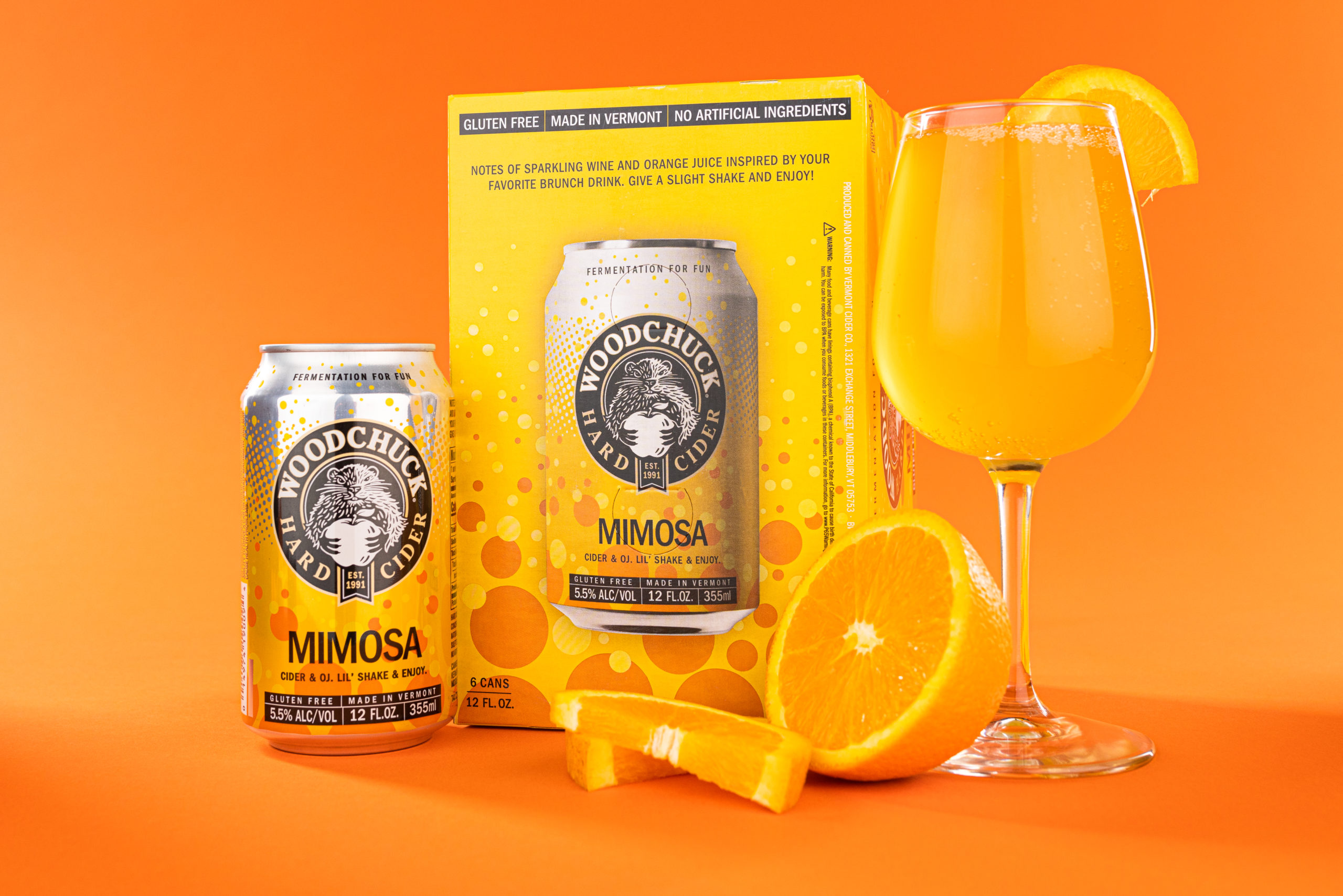 A 12 oz can and a 6 pack of Woodchuck Hard Cider Mimosa - Cider and Oj, Lil' shake and enjoy- next to a tall wine glass filled with orange mimosa Cider and a fresh orange cut in half. The 6 pack reads "Notes of sparkling wine and orange juice inspired by your favorite brunch drink. Give a slight shake and enjoy!"
