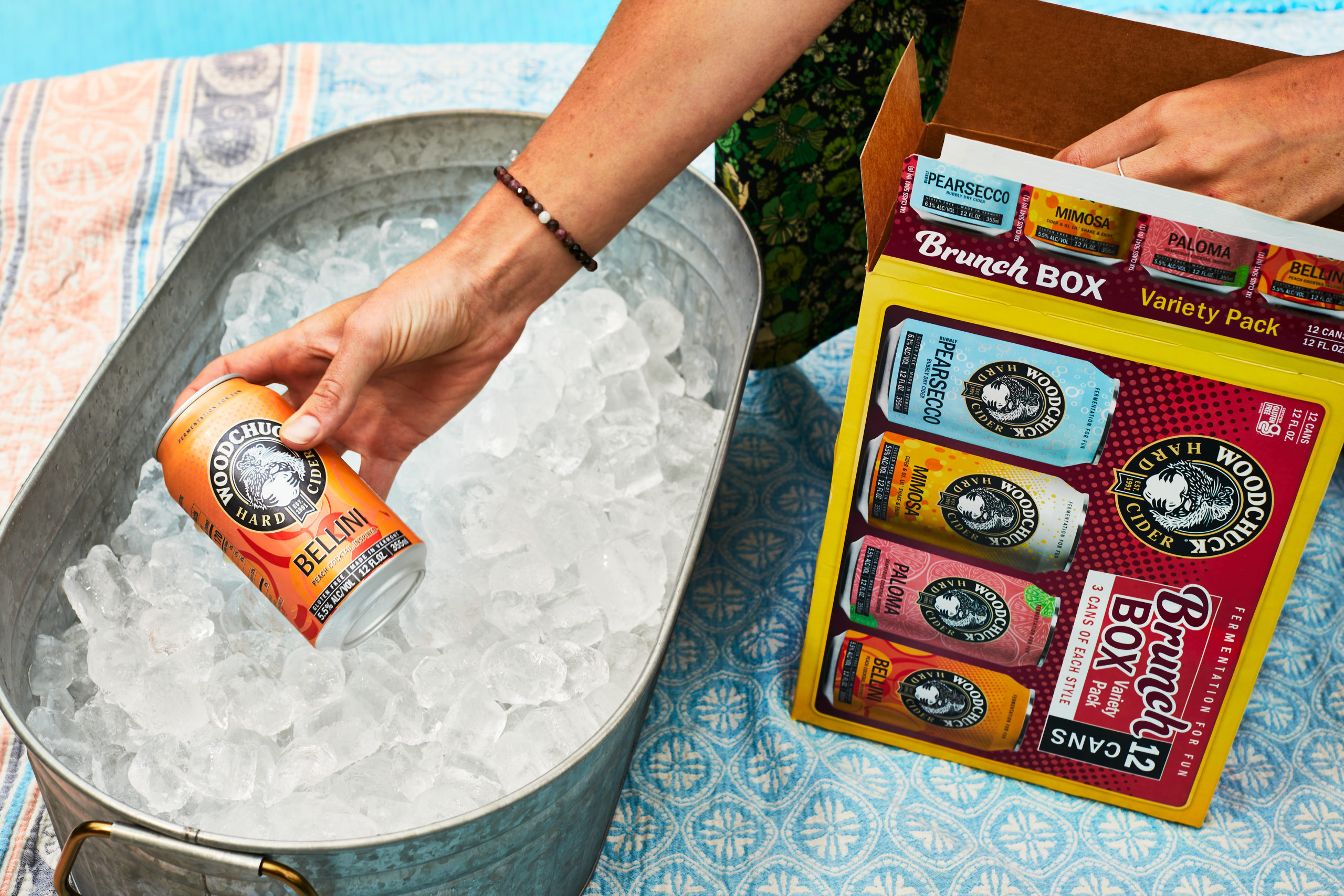 A hand placing a 12 oz can of Woodchuck Bellini Hard Seltzer in a bucket of ice while grabbing another can out of Woodchucks Brunch Box 12-can variety pack