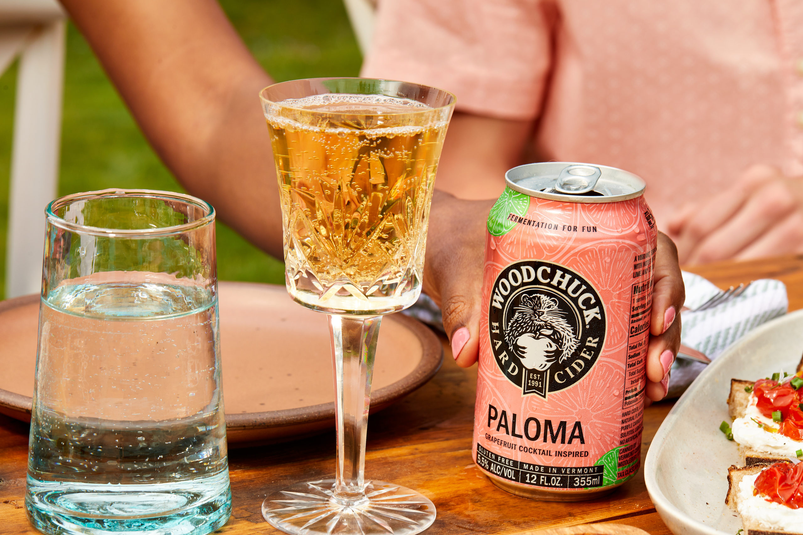 A hand holding a 12 oz can of Woodchuck Hard Ciders Paloma- grapefruit inspired cocktail - next to a champagne glass filled with sparkling Paloma Cider